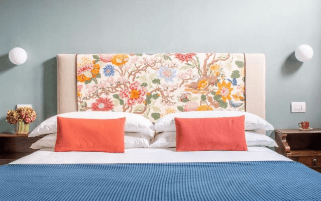 Bedroom with floral headboard, blue blanket, orange pillows, and wall sconces.